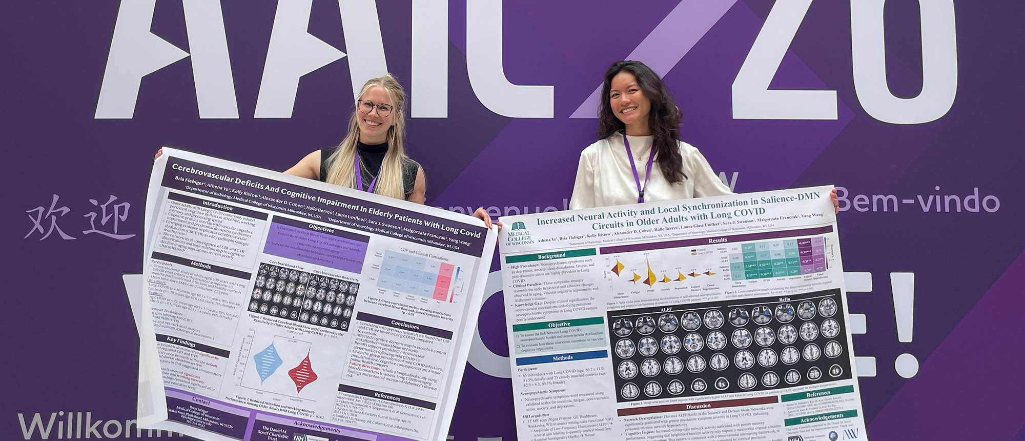 Two women holding research posters in front of conference banner