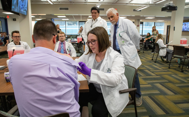 Ana Flu Clinic_Image Card Female pharmacy student administers a flu vaccine to a patient while faculty observe.