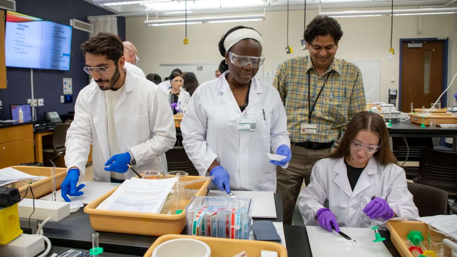Professor working with three students wearing white coats and protective eyewear in the compounding lab.