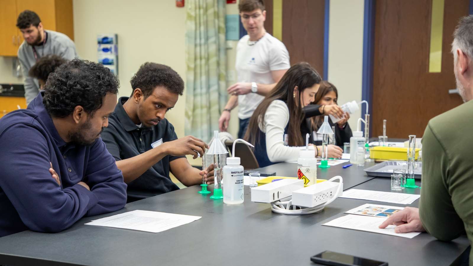 Pairs of people sitting at a lab table measuring liquid in a graduated cylinder.