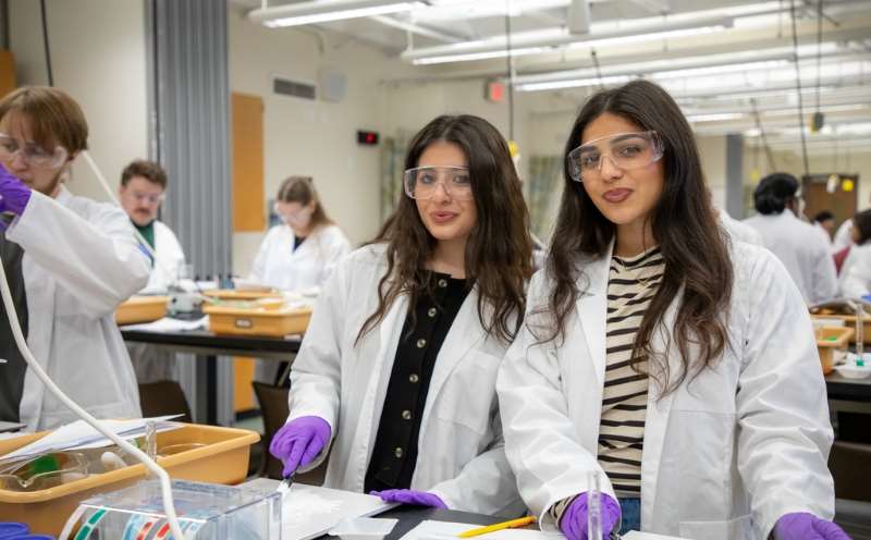 Two women wearing white coats and safety glasses in the lab classroom.