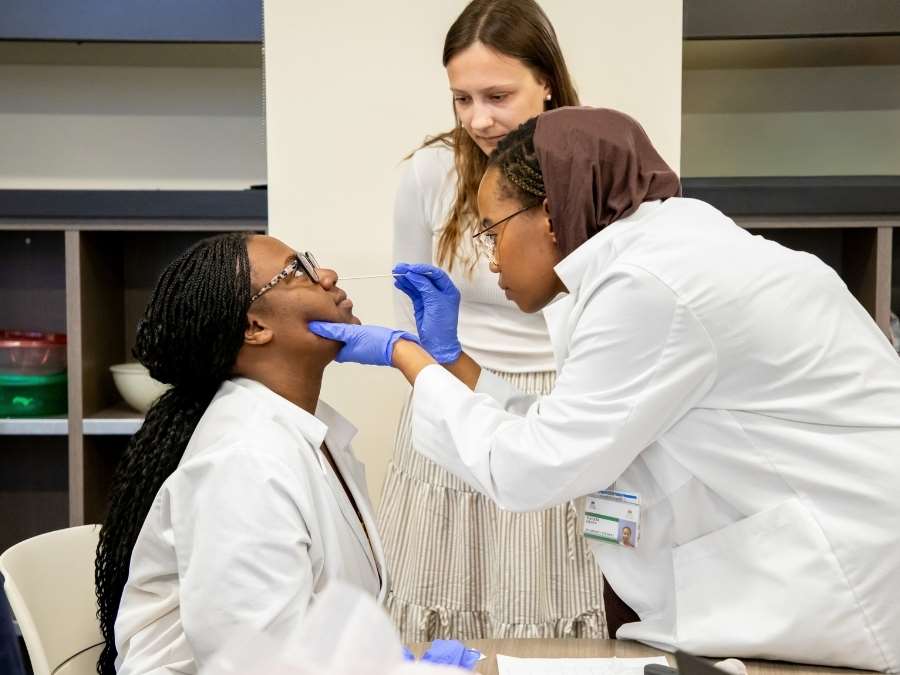 Woman in white lab coat and gloves swabs nose of PharmD classmate while instructor observes.