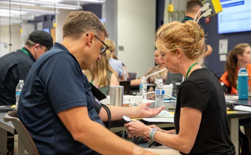 Adv Physical Assessment_Image Card Woman reads blood pressure of man wearing blood pressure cuff.