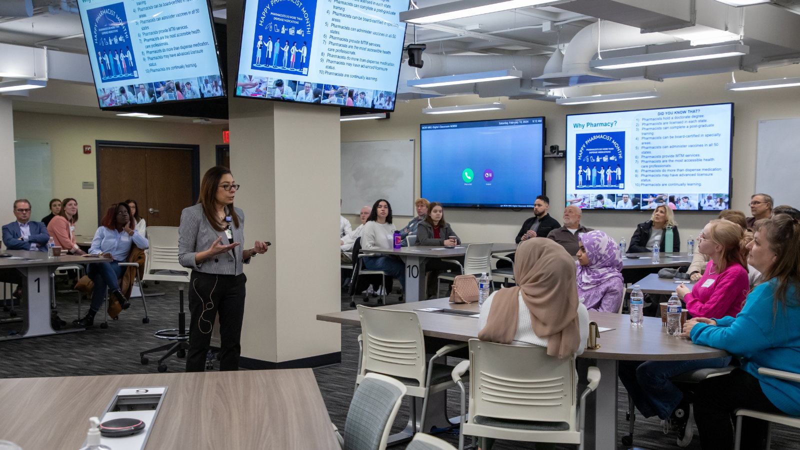 A professor speaks to a group of students sitting at tables in a classroom.