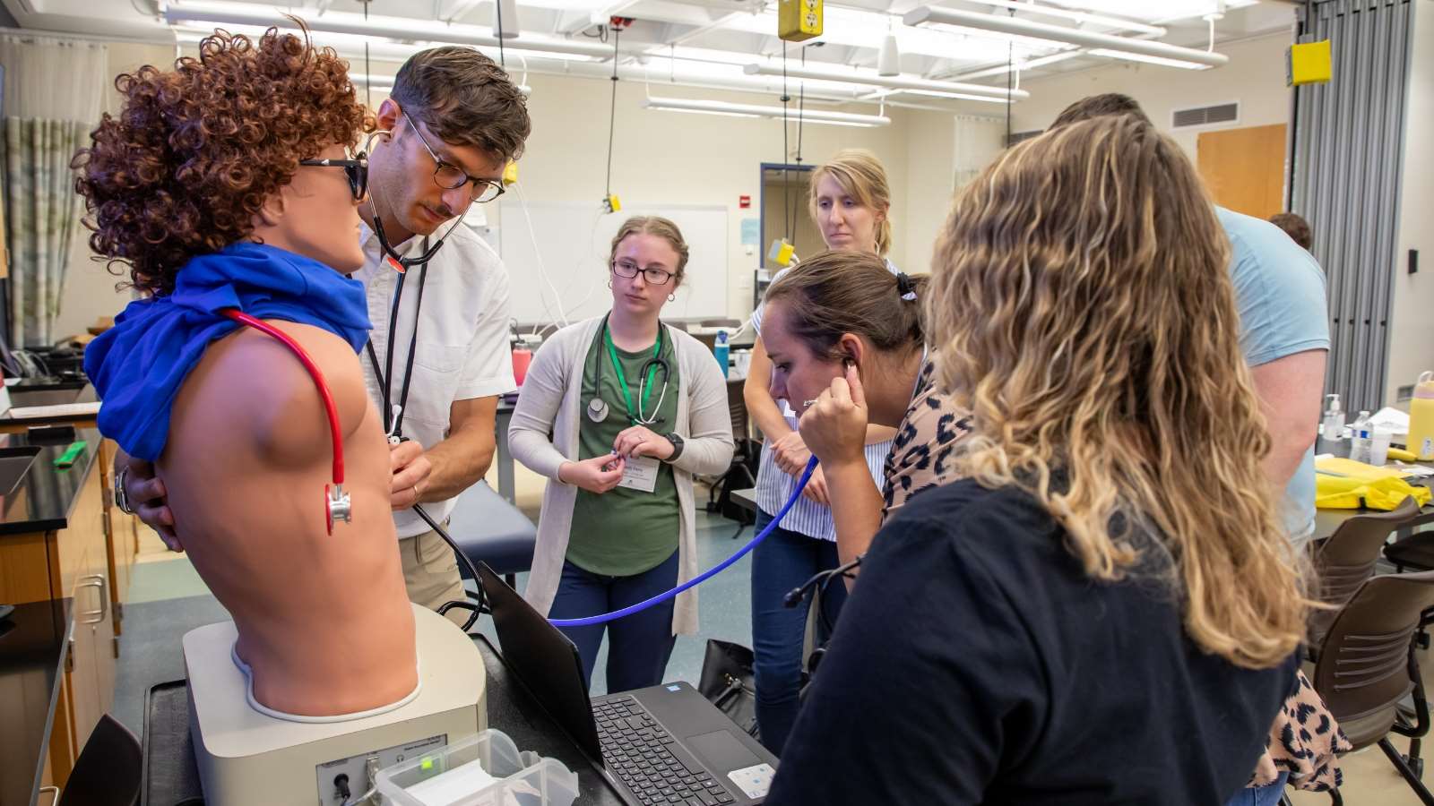 Man using a stethoscope to listen to a mannikin's heart sounds while a group of students watches.