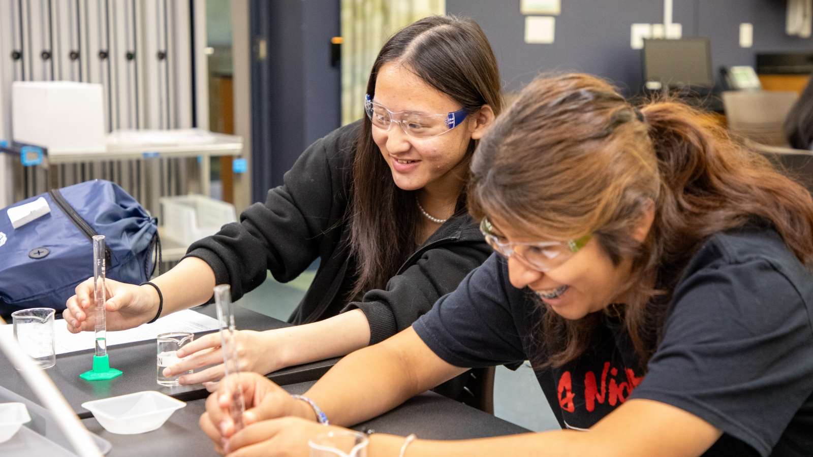 Pre-Pharmacy Scholars Lab_Image Text Split Two female students wearing safety glasses measure liquid in graduated cylinders.