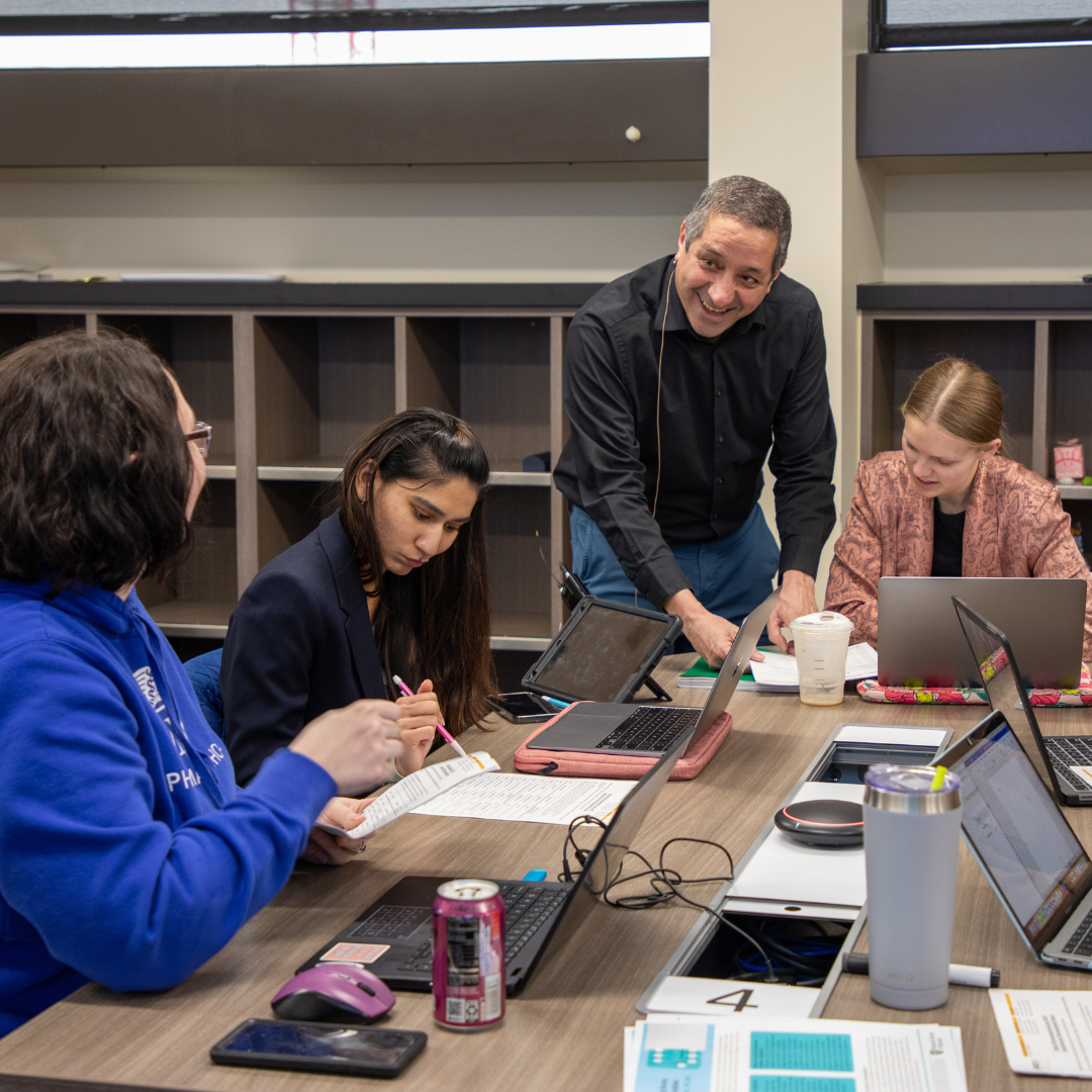 Small Class Size Professor talks with three pharmacy students sitting around a table.