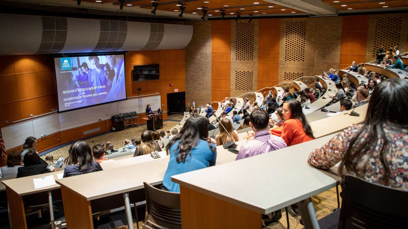 Research and Scholarship Forum_Image Text Split Group of people in a large lecture hall gather to hear a research presentation.