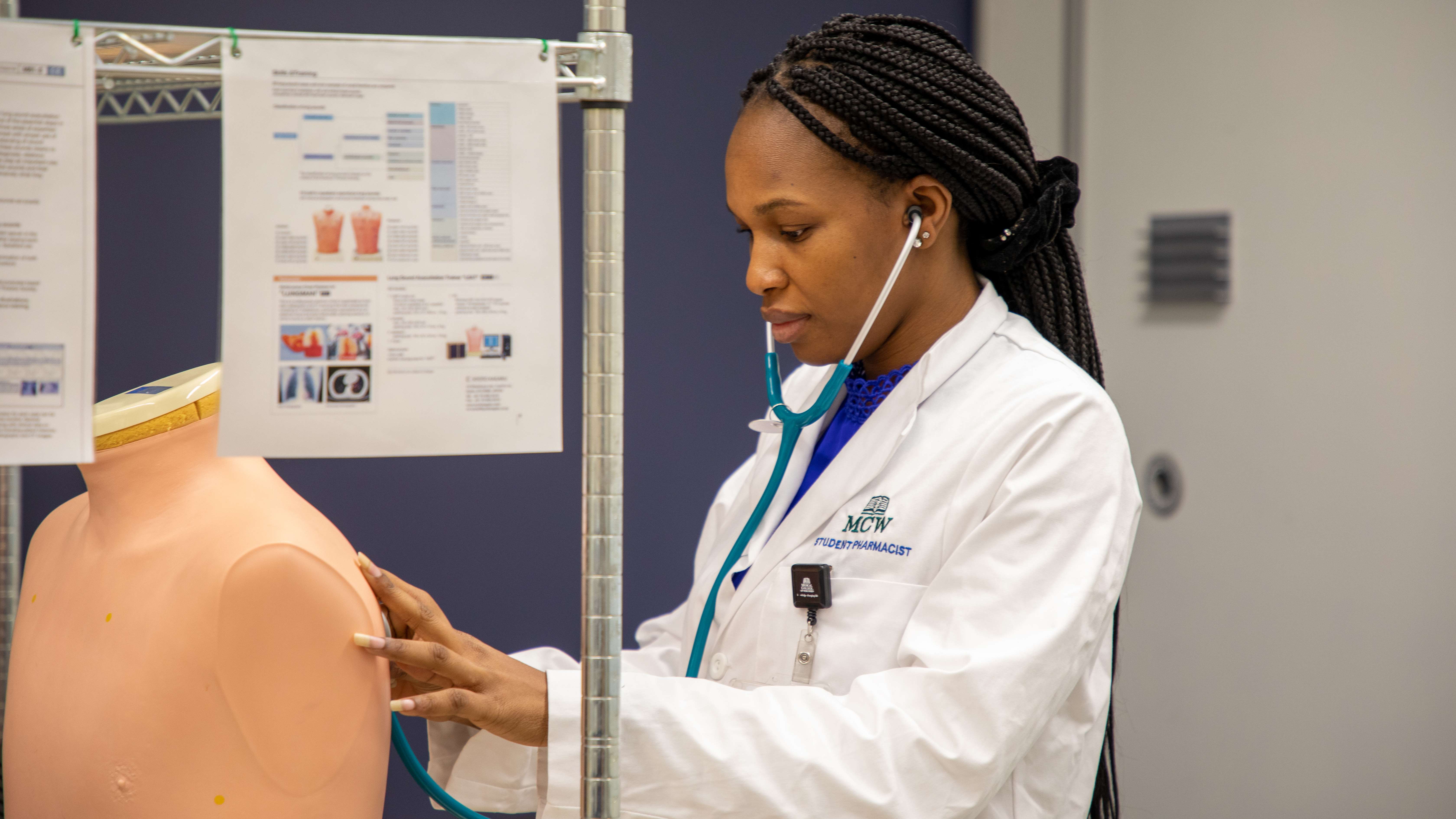 Girl in white lab coat uses a stethoscope to listen for lung sounds on a manikin.