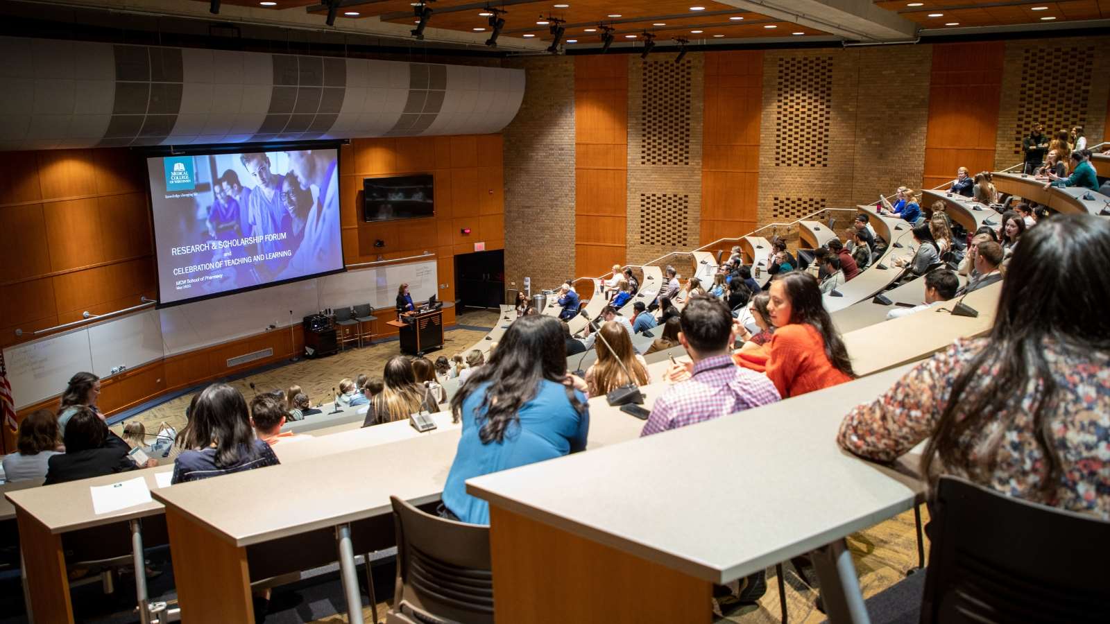 Group of people in a large lecture hall gather to hear a research presentation. 