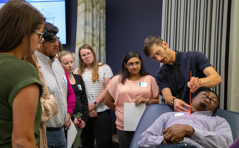 Professor shows a group of students how to perform cardiovascular assessment on a male sitting on a medical table.